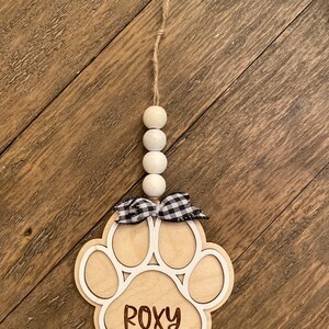 May include: A wooden paw print ornament with the name "ROXY" engraved on it. The ornament is decorated with a black and white checkered ribbon and a string of white beads.