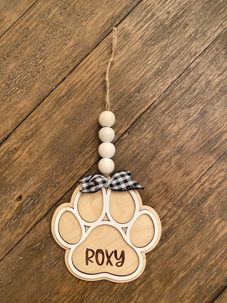 May include: A wooden paw print ornament with the name "ROXY" engraved on it. The ornament is decorated with a black and white checkered ribbon and a string of white beads.