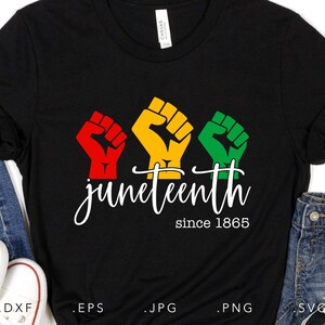 以下が含まれることがあります： 赤、黄、緑の3つの拳が描かれた黒いTシャツ。白い筆記体で「juneteenth」と「since 1865」の文字がプリントされています。ジーンズと白いスニーカーと一緒に表示されています。