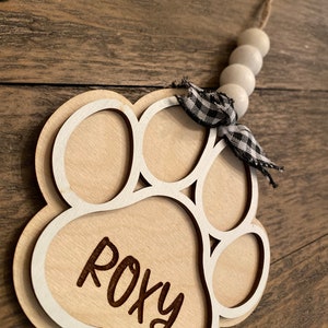 May include: A wooden paw print ornament with the name "Roxy" engraved on it. The ornament is white with a black and white checkered ribbon and a string of white beads.