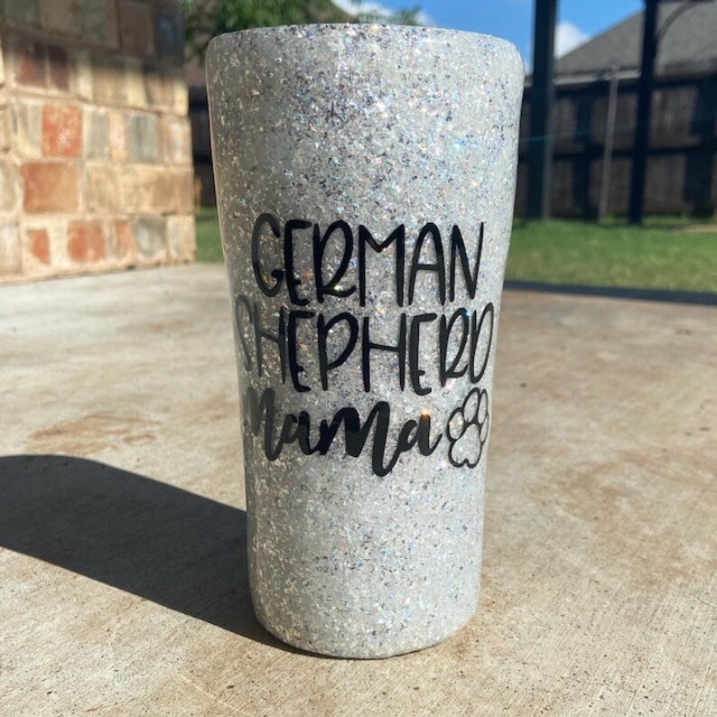 May include: A white glitter tumbler with a black design that says "German Shepherd Mama" with a paw print.