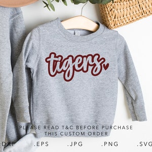 May include: Gray sweatshirt with a maroon "tigers" design and a small heart. The text "PLEASE READ T&C BEFORE PURCHASE THIS CUSTOM ORDER" is printed below the design. The sweatshirt is on a hanger.