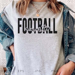 May include: A light gray t-shirt with the words "FOOTBALL aunty" printed in black. The word "FOOTBALL" is in a bold, block font, and "aunty" is in a cursive script. The shirt is being worn with a denim jacket.
