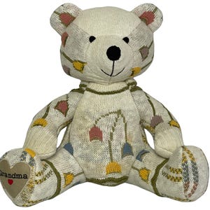 May include: A cream-coloured knitted teddy bear with a black nose and embroidered details. The bear has a heart-shaped patch on its foot with the word "Grandma" and a red heart. The bear is decorated with colourful geometric patterns.