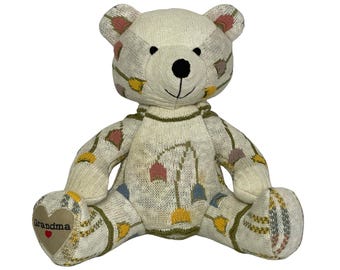 May include: A cream-colored knitted teddy bear with a black nose and embroidered details. The bear has a heart-shaped patch on its foot with the word "Grandma" and a red heart. The bear is decorated with colorful geometric patterns.