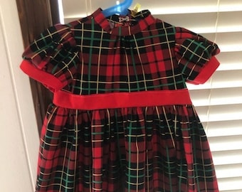 Könnte beinhalten: Ein rot-schwarz-grünes kariertes Kleid mit kurzen Ärmeln und einem roten Bund. Das Kleid hängt an einem blauen Kleiderbügel vor einem Fenster mit weißen Jalousien. Das Kleid hat einen hohen Ausschnitt.