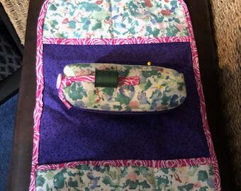 Puede incluir: Un proyecto de costura de tela morada y blanca con estampado floral. El proyecto tiene un bolsillo con un alfiletero dentro. El alfiletero está lleno de alfileres y una bobina de hilo.