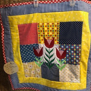 May include: A yellow, red, blue, and white patchwork quilt with a floral design. The quilt is made of squares of different fabrics and has a red and white gingham border. The quilt is hanging on a wall.