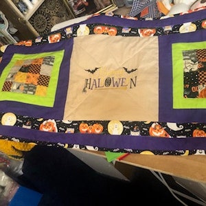 May include: A purple and green Halloween table runner with a patchwork design and the word "HALLOWEEN" embroidered in the center.
