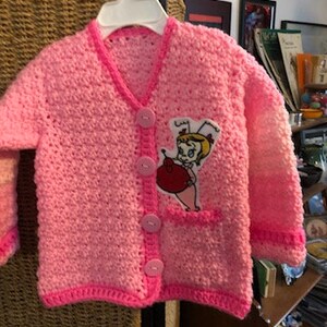 Könnte beinhalten: Rosa gestrickte Strickjacke mit einem Comic-Charakter-Aufnäher. Der Aufnäher zeigt ein Mädchen in einem roten Kleid und einem weißen Hut. Die Strickjacke hat einen Knopfverschluss und eine Tasche auf der linken Seite.
