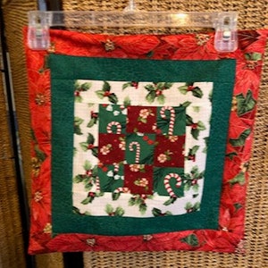 Könnte beinhalten: Ein quadratisches, gestepptes Weihnachtskissen mit rotem Poinsettia-Rand. Die Mitte zeigt ein Patchwork-Design mit Zuckerstangen, Stechpalmen und roten und grünen Quadraten. Es hängt an einem transparenten Kunststoffbügel.
