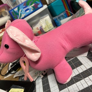 May include: A pink stuffed dachshund dog with floppy ears and a pink tail. The dog has black button eyes and a pink ribbon attached to its neck. The ears have white polka dots. The dog is lying on a cutting mat.