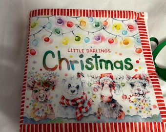Pode incluir: Um livro de Natal de tecido com uma borda listrada vermelha e branca. A capa apresenta as palavras "Little Darlings Christmas" e ilustrações de animais festivos com decorações e luzes de Natal.