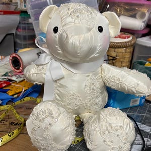 Può includere: Un orsacchiotto color crema in raso, decorato con pizzo e perle. Ha un nastro bianco e guance rosee. Le zampe e il corpo dell'orso sono impreziositi da pizzo, creando un design delicato. Un oggetto decorativo affascinante.