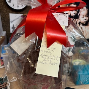 May include: A gift basket wrapped in clear cellophane with a red ribbon. The basket contains a variety of items, including a ceramic cookie tray, a spatula, a handmade quilted table runner, and a house-shaped basket. The basket is labeled with the text "Handmade Quilted Table RUNNER by ANNIE B House Bastet".