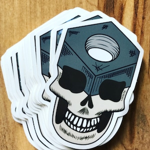 May include: A stack of white stickers with a black and white skull design. The skull is wearing a gray metal nut on its head.