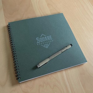 May include: A green spiral-bound notebook with the text "SQUARE SCOREBOOK 60" printed on the cover. A beige pen rests on the notebook. The notebook and pen are on a light wood surface.