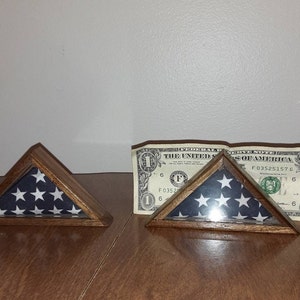 May include: Two wooden shadow boxes with glass fronts displaying folded American flags. The boxes are triangular in shape and have a dark wood finish. A dollar bill is visible behind the right box.