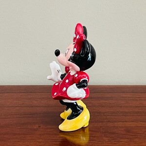 May include: A ceramic figurine of Minnie Mouse, featuring a red dress with white polka dots, a matching bow, and yellow shoes. The figure has black ears and a black nose, with white gloves. The figurine is posed with one hand raised.