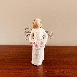 May include: A carved figurine of an angel in a white dress, holding a bouquet of pink flowers. The angel has blonde hair and wire wings. The figurine is displayed on a wooden surface.