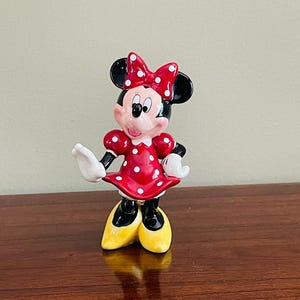 May include: A ceramic Minnie Mouse figurine. The figure is wearing a red dress with white polka dots, a matching bow, and yellow shoes. The figurine has black ears, black gloves, and a smiling expression.