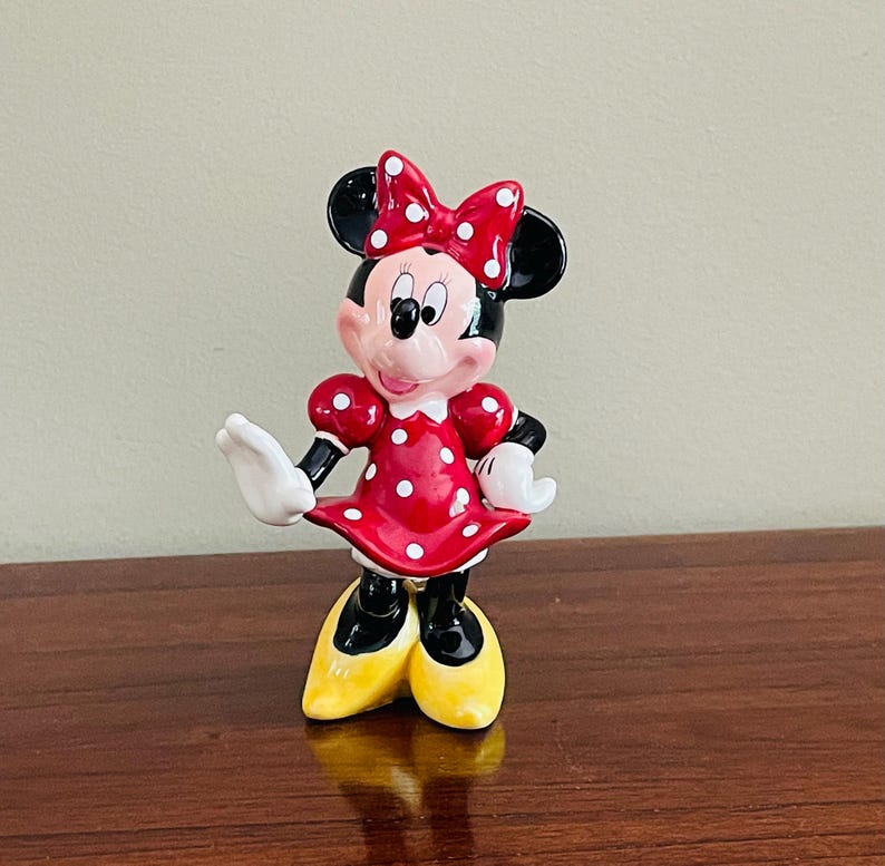 May include: A ceramic Minnie Mouse figurine. The figure is wearing a red dress with white polka dots, a matching bow, and yellow shoes. The figurine has black ears, black gloves, and a smiling expression.