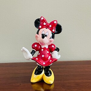 May include: A ceramic Minnie Mouse figurine. Minnie is wearing a red dress with white polka dots, a matching bow, and yellow shoes. Her black ears and black gloves are also visible. The figurine is standing on a wooden surface.