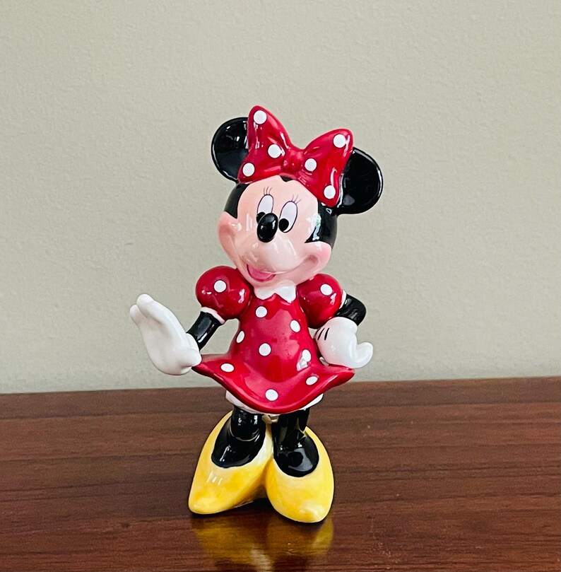 May include: A ceramic Minnie Mouse figurine. Minnie is wearing a red dress with white polka dots, a matching bow, and yellow shoes. Her black ears and black gloves are also visible. The figurine is standing on a wooden surface.