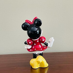 May include: A ceramic Minnie Mouse figurine, viewed from the back, with a red dress with white polka dots, a red bow, and white gloves. The figurine is standing on a yellow base.