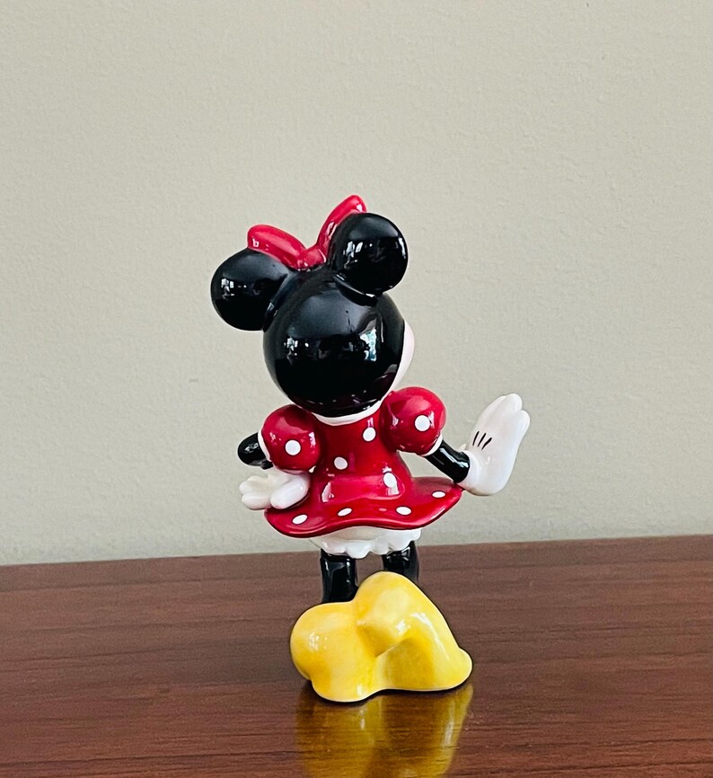 May include: A ceramic Minnie Mouse figurine, viewed from the back, with a red dress with white polka dots, a red bow, and white gloves. The figurine is standing on a yellow base.