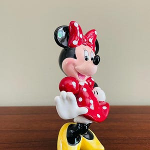 May include: A ceramic Minnie Mouse figurine. Minnie is wearing a red dress with white polka dots and a matching bow. Her shoes are yellow, and her gloves are white. The figurine has black ears and a pink face.