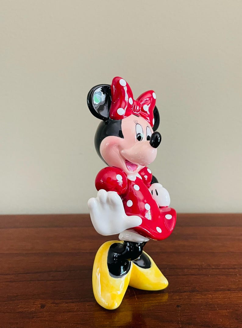 May include: A ceramic Minnie Mouse figurine. Minnie is wearing a red dress with white polka dots and a matching bow. Her shoes are yellow, and her gloves are white. The figurine has black ears and a pink face.
