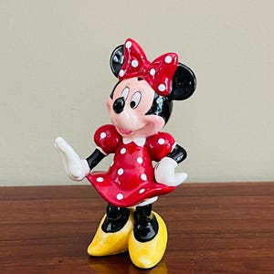 May include: A ceramic figurine of Minnie Mouse, a classic Disney character. She wears a red dress with white polka dots, a matching bow, and yellow shoes. Her black ears and gloves are also visible.