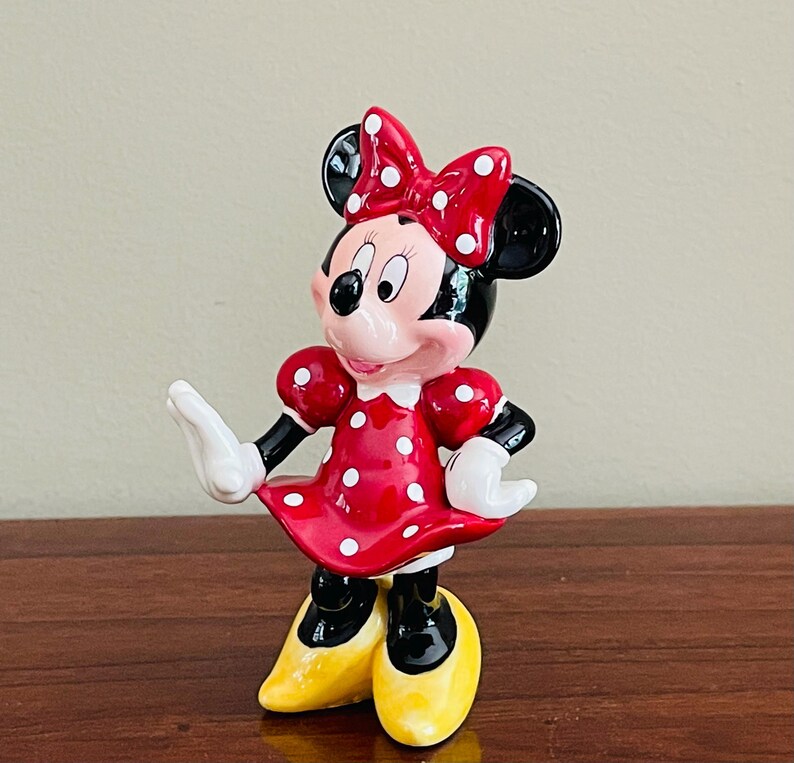 May include: A ceramic figurine of Minnie Mouse, a classic Disney character. She wears a red dress with white polka dots, a matching bow, and yellow shoes. Her black ears and gloves are also visible.