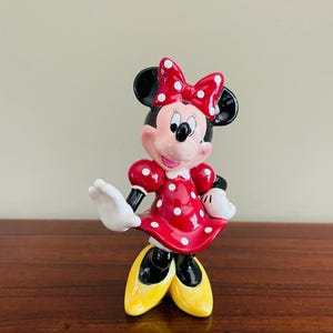 May include: A ceramic Minnie Mouse figurine. The figure is wearing a red dress with white polka dots, a matching bow, and yellow shoes. The figurine has black ears, a pink face, and white gloves. The figure is posed with one arm raised.