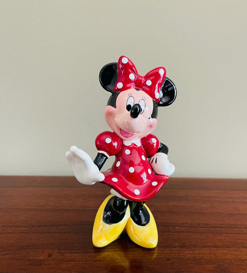 May include: A ceramic Minnie Mouse figurine. The figure is wearing a red dress with white polka dots, a matching bow, and yellow shoes. The figurine has black ears, a pink face, and white gloves. The figure is posed with one arm raised.
