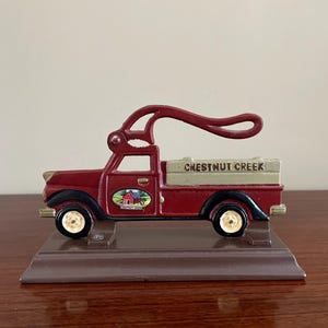 May include: A red and black cast iron nutcracker shaped like a vintage pickup truck. The truck bed is beige with the text CHESTNUT CREEK printed on it. A small oval decal on the door shows a red barn. The truck sits on a brown rectangular base.