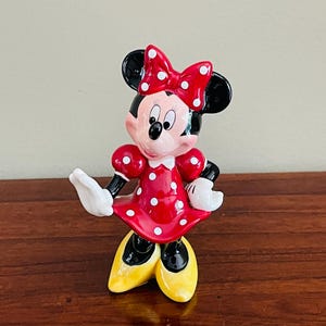 May include: A Minnie Mouse figurine in a classic pose. The figure is wearing a red dress with white polka dots, a matching bow, and yellow shoes. The figurine has black ears, a black nose, and white gloves.