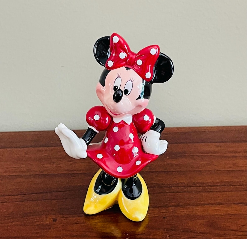 May include: A Minnie Mouse figurine in a classic pose. The figure is wearing a red dress with white polka dots, a matching bow, and yellow shoes. The figurine has black ears, a black nose, and white gloves.