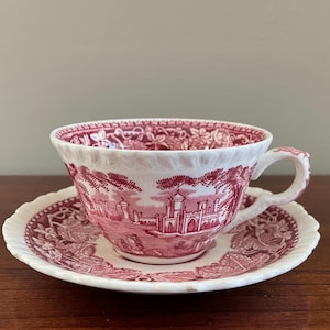 May include: A white teacup and saucer set with a red and white toile pattern. The teacup has a handle and sits on a matching saucer. The design features a detailed landscape scene. The set is on a wooden surface.