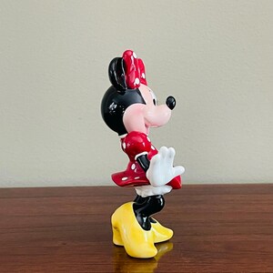 May include: A ceramic figurine of Minnie Mouse, featuring a black head and ears, a red dress with white polka dots, and yellow shoes. The figurine has a red bow with white polka dots and white gloves. The figure is posed in a playful stance.