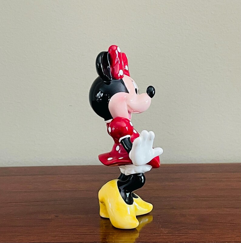 May include: A ceramic figurine of Minnie Mouse, featuring a black head and ears, a red dress with white polka dots, and yellow shoes. The figurine has a red bow with white polka dots and white gloves. The figure is posed in a playful stance.