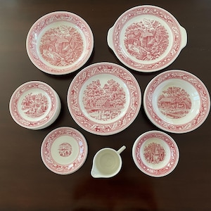 May include: A collection of white ceramic dinnerware featuring a red transferware pattern of rural landscapes and buildings. The set includes various plates, bowls, and a small creamer pitcher, all decorated with intricate red floral borders.