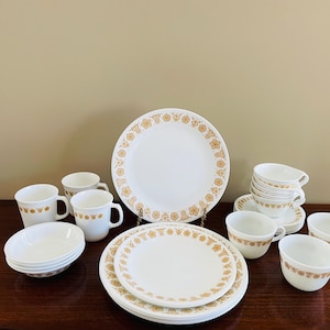 May include: A complete vintage white dinnerware set with a gold floral pattern. The set includes plates, bowls, cups, and saucers. The plates and cups have a gold floral border. The set is arranged on a dark wooden surface.