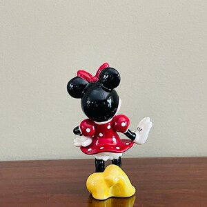 May include: A ceramic Minnie Mouse figurine, viewed from the back, with a red dress with white polka dots, a red bow, and black ears. The figurine has white gloves and black shoes. A yellow object is in front of the figurine.