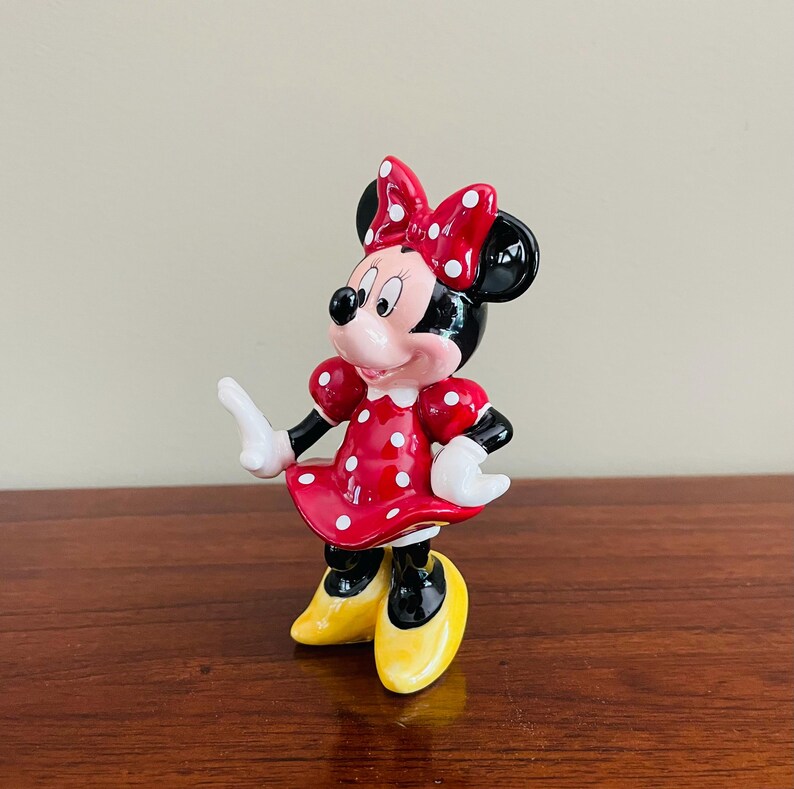 May include: A Minnie Mouse figurine, featuring a red dress with white polka dots, a matching bow, and yellow shoes. The black-eared character has white gloves and a cheerful expression. The figurine is standing on a wooden surface.