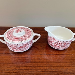May include: A white ceramic sugar bowl with a lid and a matching creamer, both featuring a detailed red transferware pattern of landscapes and floral motifs. The sugar bowl has two side handles, and the creamer has a pouring spout and one handle.