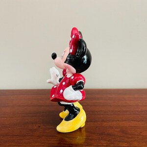 May include: A Minnie Mouse figurine, featuring a red dress with white polka dots, a matching bow, and yellow shoes. The figurine has a black head and ears, and white gloves. The classic Disney character is posed in a side view.