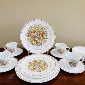May include: A vintage white dinnerware set with floral designs. The set includes plates, saucers, cups, and bowls. The plates and bowls have a brown trim. The floral pattern features orange, yellow, and green flowers.