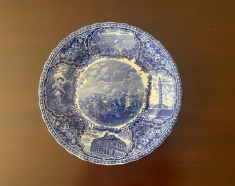 May include: A round ceramic plate with a scalloped edge, featuring a blue and white transferware pattern. The center depicts a battle scene, surrounded by vignettes of historical landmarks and figures, including text labels like New Orleans.